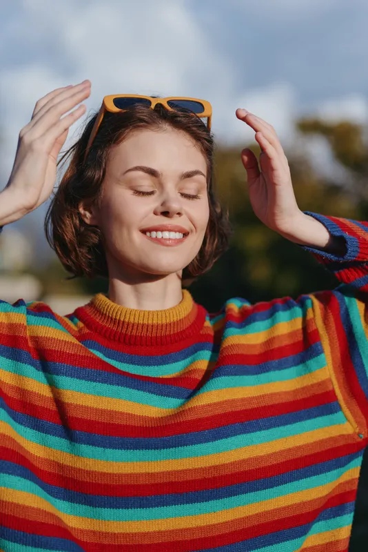 Woman portrait smiling with eyes closed wearing sunglasses on head and colorful sweater outdoors in sunlight. Happy young woman enjoying warm day and relaxed moment.