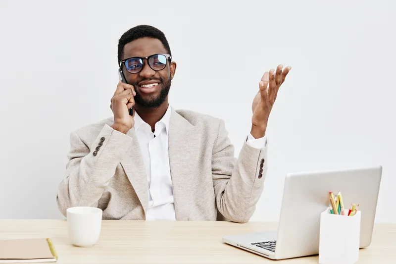 Smiling man with glasses talking on mobile phone while sitting at desk with laptop, white background, casual business attire, modern office environment, cheerful mood, professional appearance.
