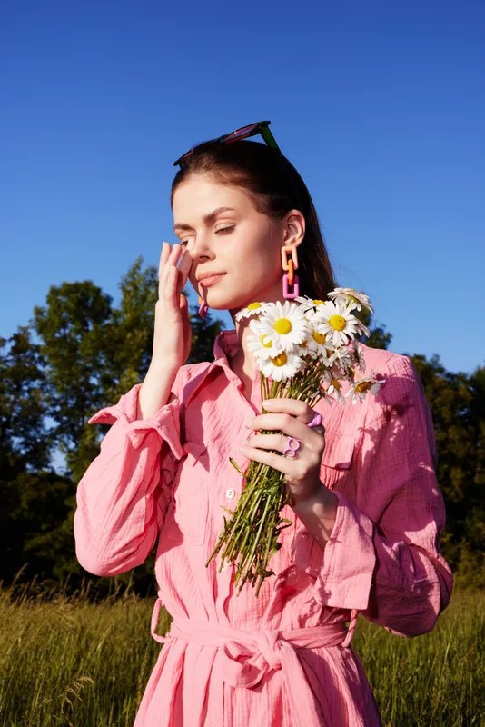 Young model in a pink dress holding daisies, showcasing joy and tranquility in a sunny field with vibrant greenery and blue sky