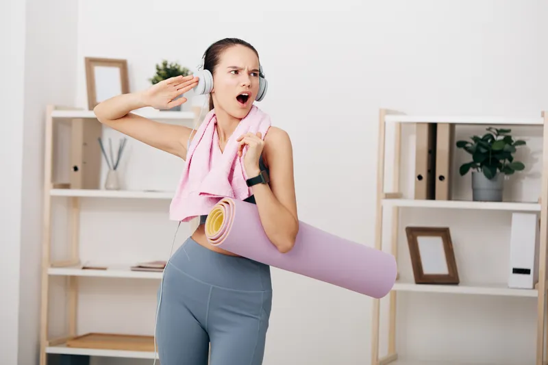 A woman in fitness attire stands in a bright home studio with headphones, a rolled yoga mat, towel draped over shoulder, ready for a workout and stretch.