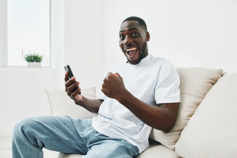 Happy young man celebrating success while using his smartphone in a bright living room, showing excitement with casual attire