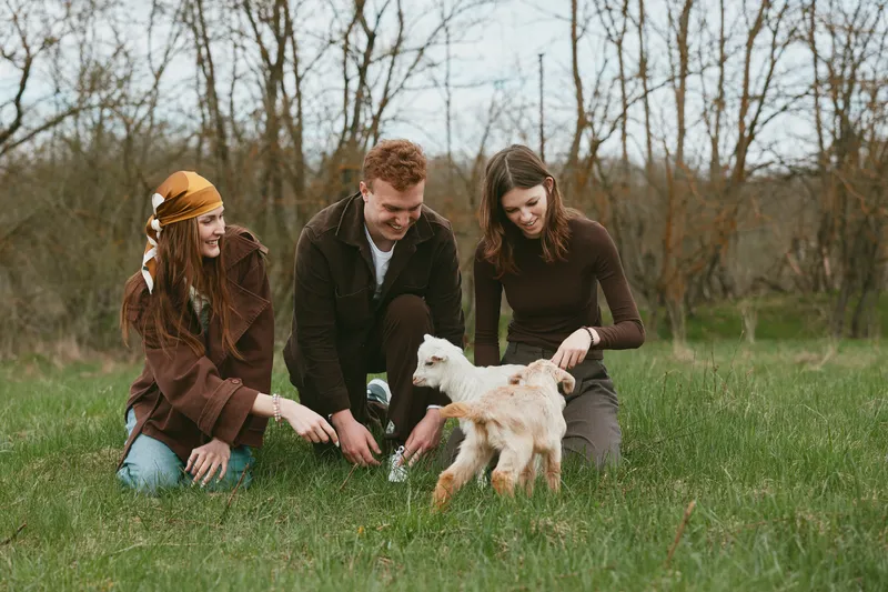 Cheerful group outdoors with a playful goat kid, laughing and interacting on a sunlit meadow, casual outfits, relaxed pose, natural colors, soft breeze, rustic trees in background, candid lifestyle