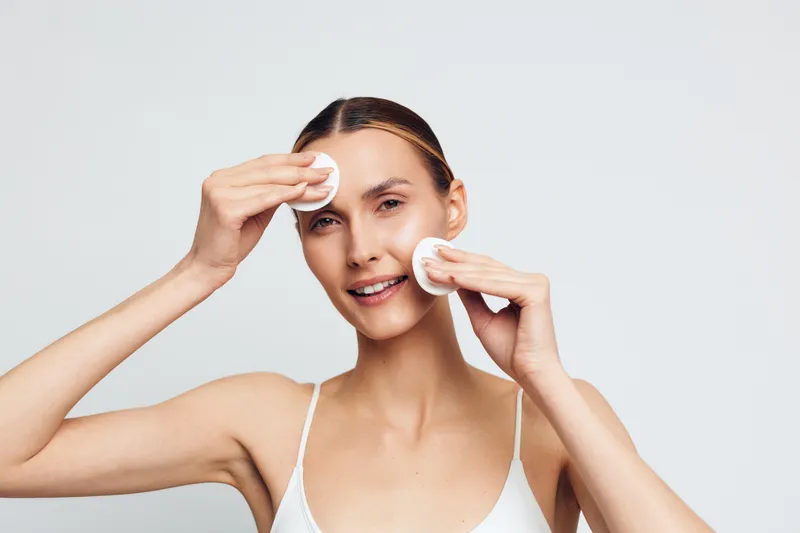 Young woman with clean skin removing makeup with cotton pads on white background. Concept of skincare, beauty routine, and facial cleansing. Soft lighting highlights natural complexion.