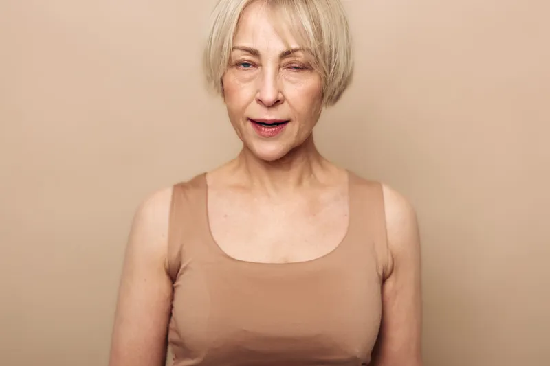 Mature woman with short blonde hair wearing beige top winks with one eye on beige background. Concept of playful expression, confidence, and natural beauty in middle age woman.
