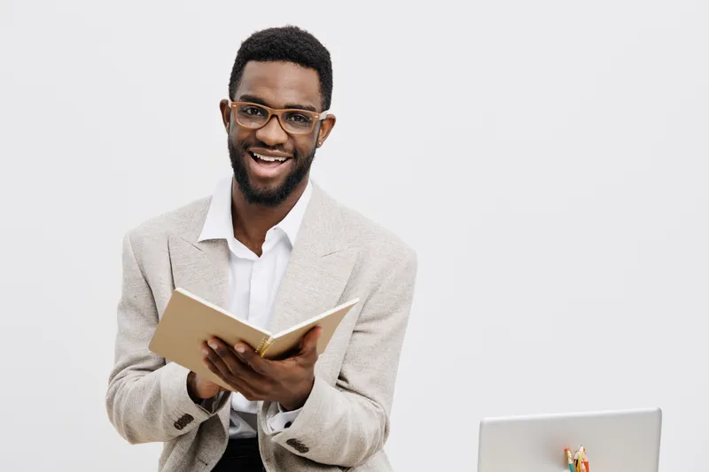 Smiling man with glasses wearing beige cardigan and white shirt reading a book while sitting at desk isolated on plain white background, professional lifestyle concept