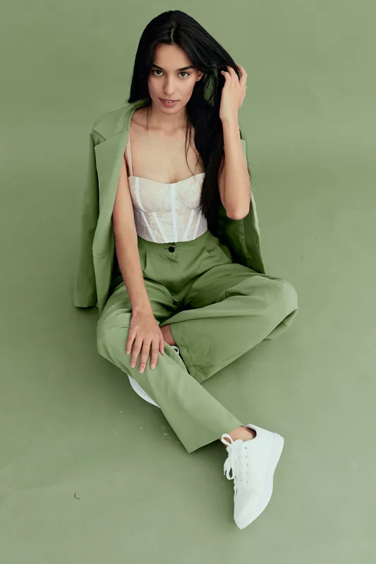 Stylish woman sitting on green floor wearing light jacket and pants with white sneakers, dark hair and confident pose in minimalistic studio setting.