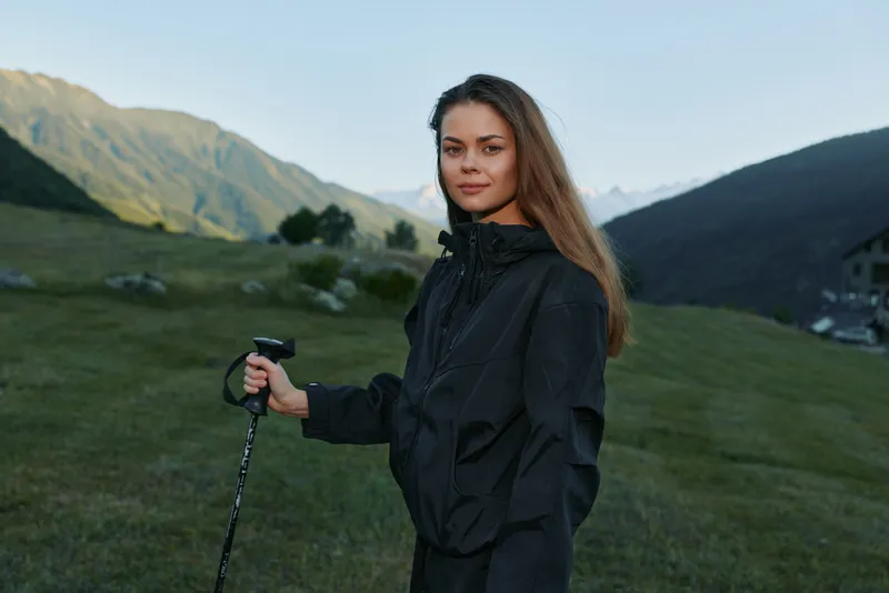 A confident hiker stands in a lush mountain meadow, gripping a trekking pole in a black rain jacket. She looks toward the distant peaks, ready for adventure.