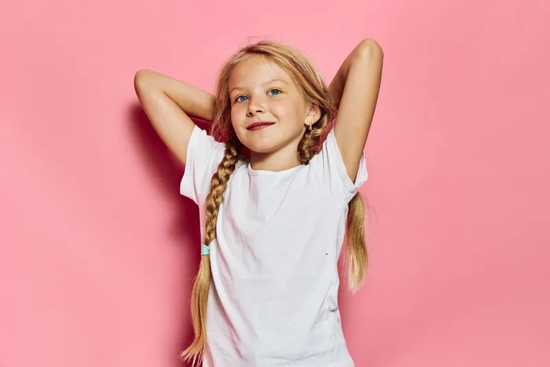 Blonde girl with braids smiles confidently with hands behind head wearing plain white t-shirt against pink background. Studio portrait. Concept of childhood, happiness, and innocence.