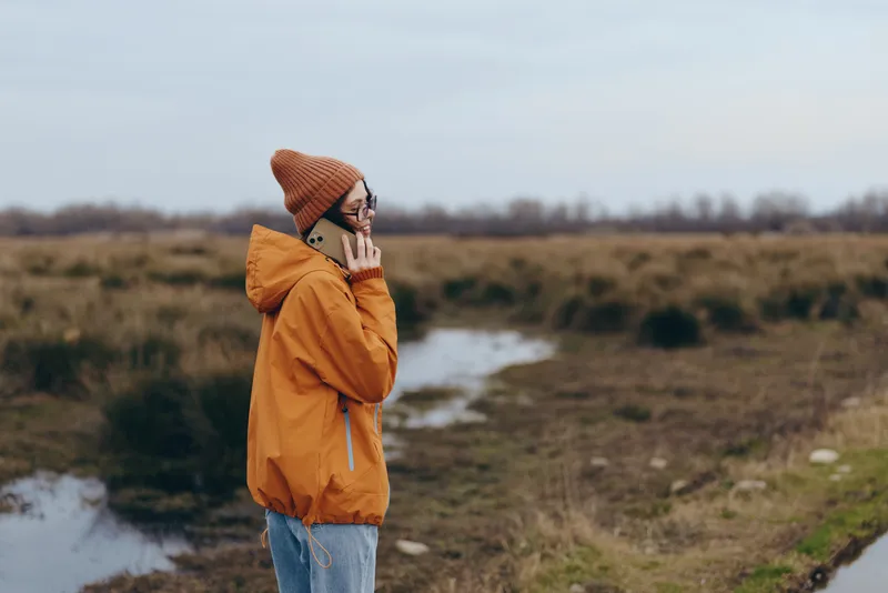 Woman lifestyle outdoors in winter jacket and beanie, countryside path, nature landscape, orange coat, smiling while holding smartphone, cloudy sky, walking slowly in peaceful rural field.