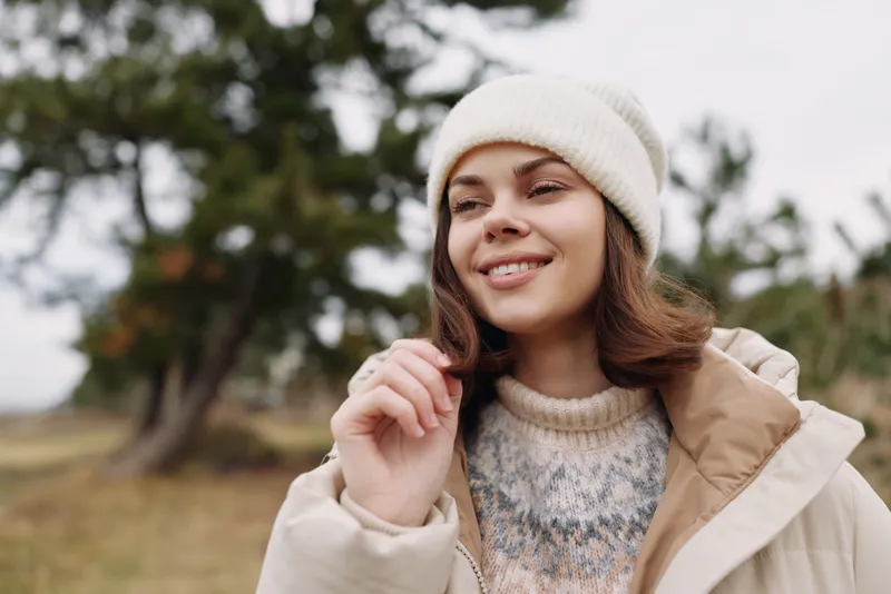 Woman portrait in outdoor setting wearing a cozy sweater and beige jacket with a knit beanie, smiling as she enjoys nature and cool weather.