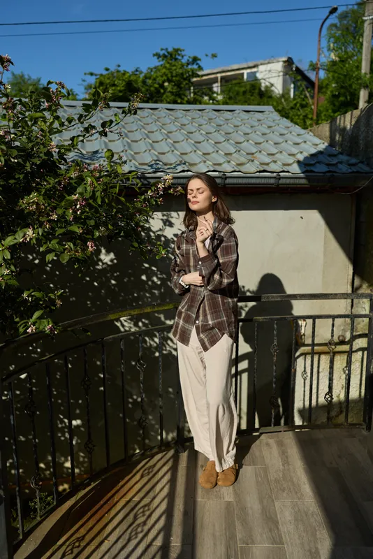 A young woman stands on a sunlit balcony wearing a plaid shirt and beige wide leg pants, eyes closed as she savors a quiet moment outdoors, while railing shadows create patterns on the concrete