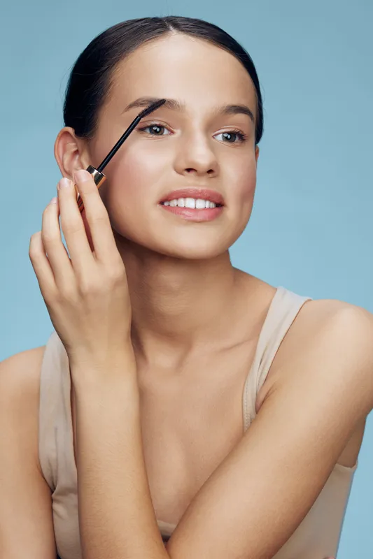 Young woman applying eyebrow gel with a natural smile, showcasing a fresh beauty look against a soft blue background, emphasizing makeup and self care