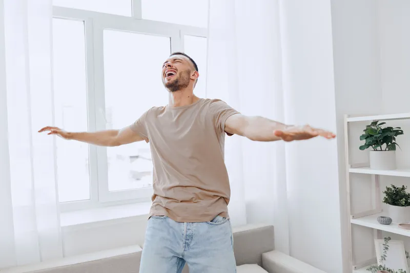 Happy man celebrating personal achievements in a bright, minimalist living space, embracing emotional freedom and self-acceptance while enjoying solitude.