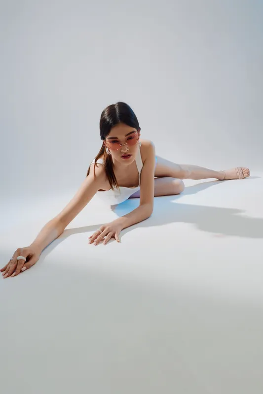 Young woman in white swimsuit and pink sunglasses poses on floor in bright studio. Concept of woman beauty fashion with minimalist style and soft shadows.