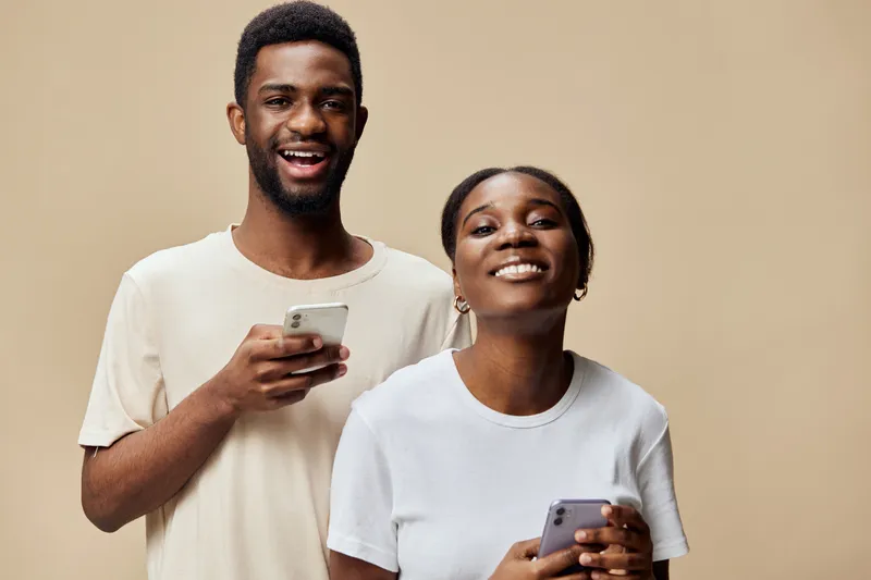 Happy young African couple with smartphones smiling in casual outfits against a beige background, representing connection and technology