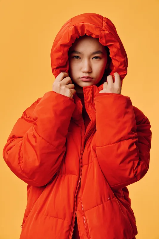 Young person in bright orange winter jacket with hood up looking serious on yellow background. Portrait of individual wearing warm coat for cold season with hands adjusting hood for protection.