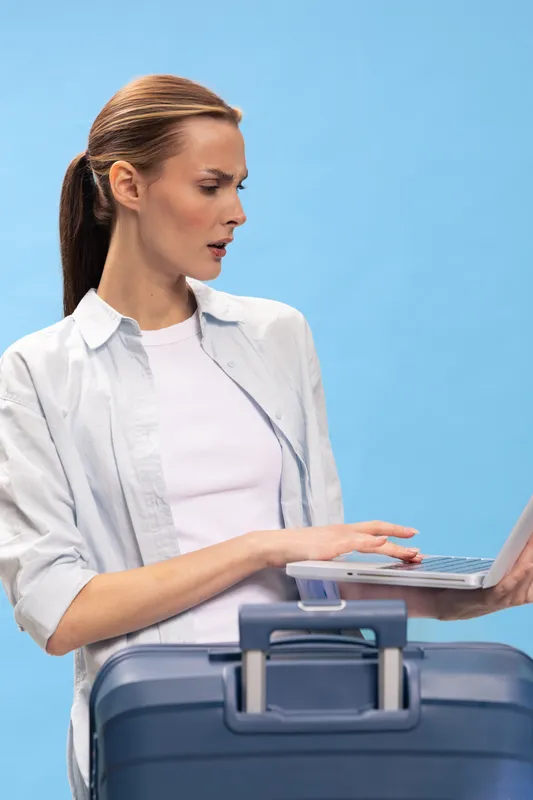 Confused young woman with ponytail looking at laptop screen, standing behind suitcase on blue background. Concept of travel planning, unexpected issues, and technology use on trip.