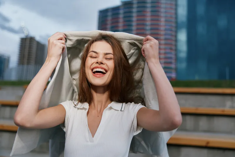 cheerful woman laughing joyfully in white top holding jacket outdoors with urban background smiling face happiness emotional expression
