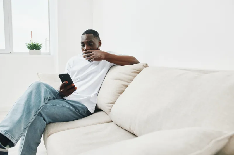 Young African American man relaxing on a light sofa while browsing on his smartphone, enjoying leisure time at home with a calm expression