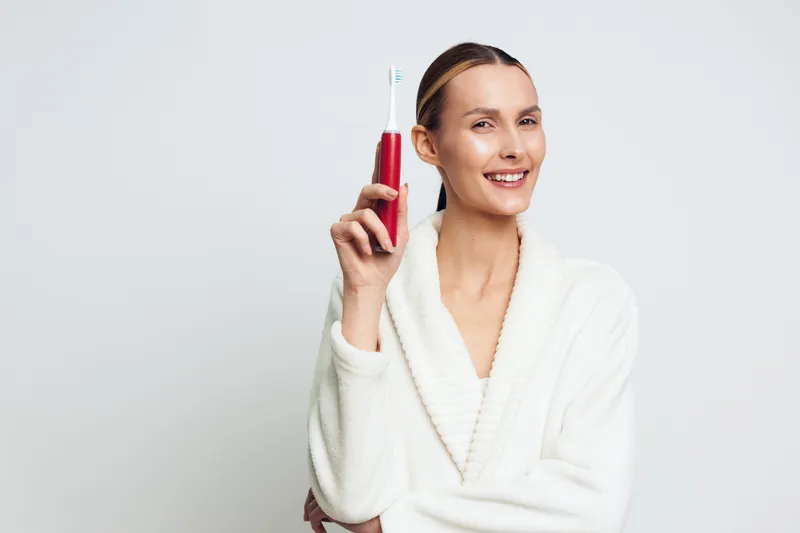 A smiling woman in a white bathrobe holds a red electric toothbrush against a plain background. The concept highlights dental care, personal hygiene, and morning routine.