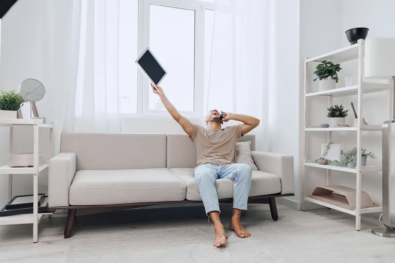 Excited man celebrating success while sitting on a stylish couch, holding a tablet high in the air. Bright, modern interior with natural light enhances the joyful atmosphere.