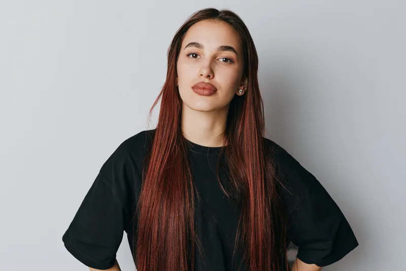 Young woman with long straight hair wearing black t shirt standing with hands on hips against plain background. Confident female portrait with natural makeup and casual style indoors.