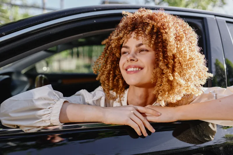 smiling woman with curly hair leaning out car window enjoying sunny day outdoors, casual clothing, natural light, positive mood, urban background