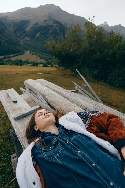 Woman lying on wooden bench in nature near mountains, portrait of relaxation in meadow with jacket and closed eyes, outdoors escape, calm scenery and peaceful rural landscape.