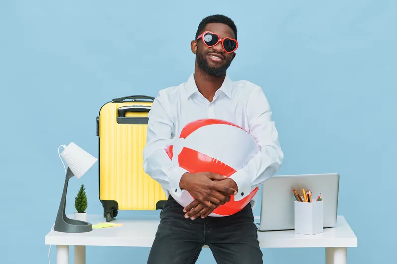 Smiling man with sunglasses holding beach ball in casual outfit standing by a colorful travel suitcase and desk with laptop on blue background. People lifestyle concept with summer vacation theme.