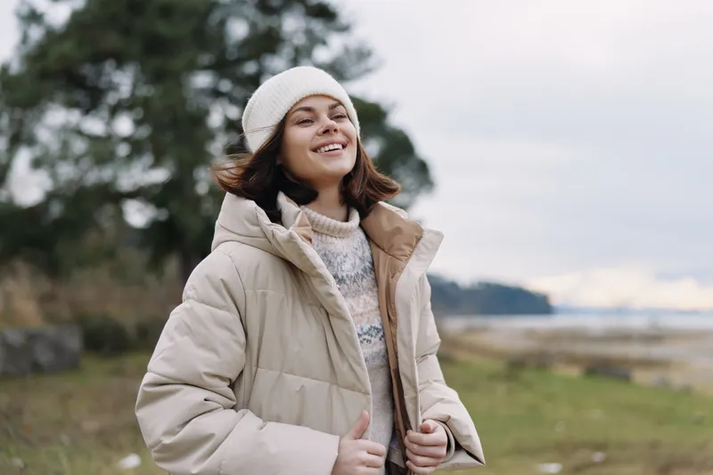 Woman wearing a beige puffer coat outdoors, smiling in a beanie and sweater, with a coastal landscape in the background for a warm casual nature portrait, soft light.