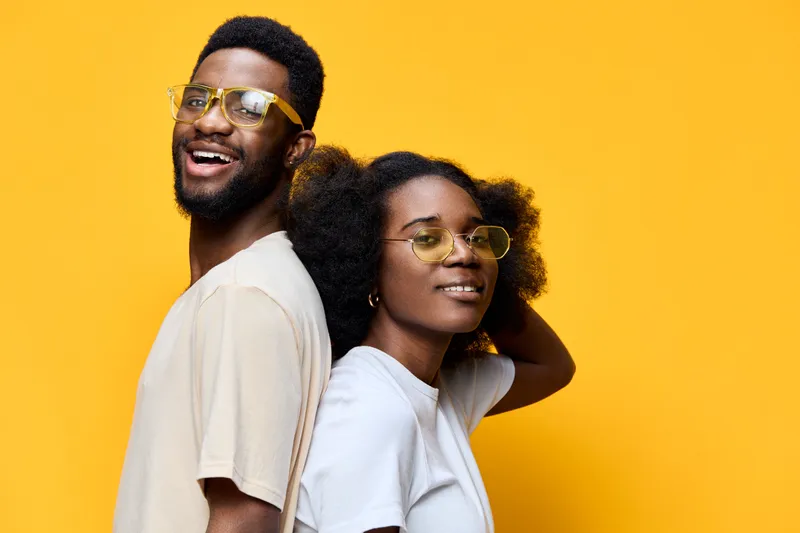 Young African couple smiling, showcasing trendy glasses against a vibrant yellow background, embodying confidence and joy in a stylish urban fashion