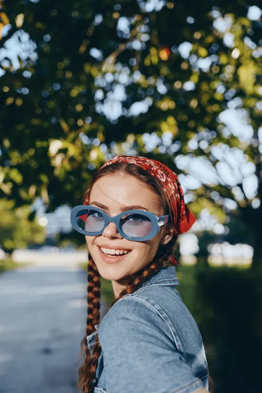 lifestyle woman in modern boho-western denim street style wearing blue sunglasses and red bandana, smiling outdoors with natural green background, film color effect