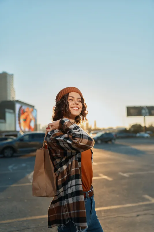 Woman smile with shopping bag in urban parking lot at golden hour, candid lifestyle portrait in beanie and casual outfit, authenticity and mindful living create emotional storytelling mood.