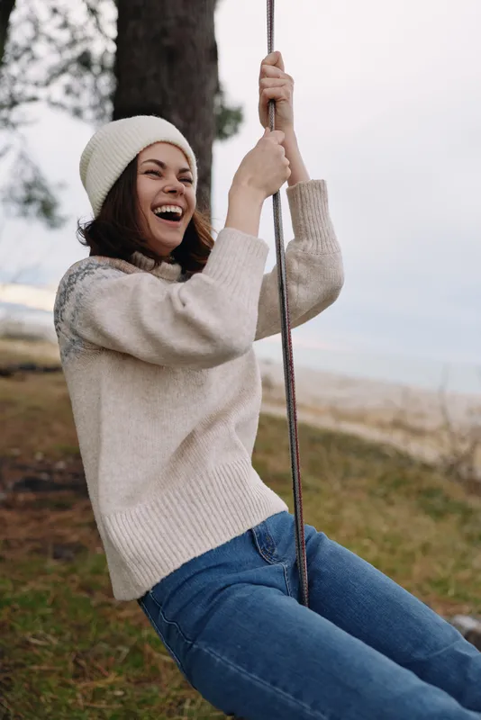 Smiling woman sits on a swing outdoors in a cozy sweater and beanie, enjoying a carefree moment in a grassy coastal park with trees and sky, conveying relaxed leisure, fashion and natural charm