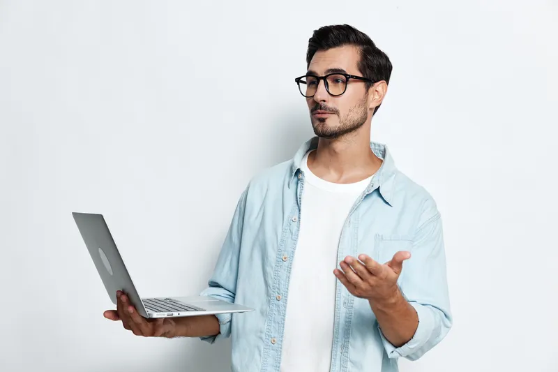 Thoughtful man wearing stylish glasses holds laptop and gestures with hand, expressing ideas or discussing work while standing against a plain light background.