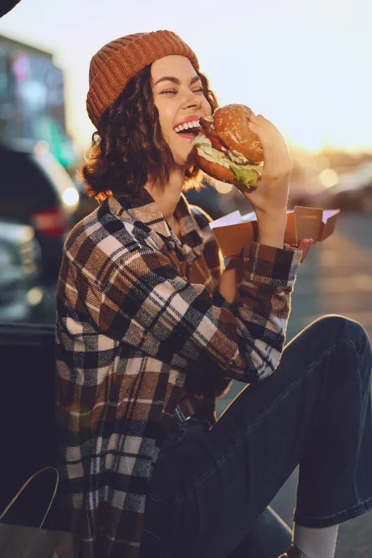 Woman with beanie and plaid shirt sits outdoor in urban street, smiling while eating a burger in sunlight. Authenticity, candid lifestyle, golden hour glow, mindful living, emotional storytelling.