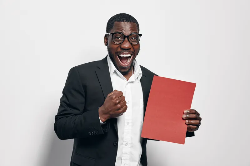 African American man with dark skin wearing glasses and suit holding red folder celebrating success with excited expression on white background.