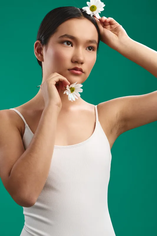 asian woman wearing a white tank top stands against a green studio backdrop, holding flowers near her face with a calm, thoughtful expression and a subtle, natural mood.
