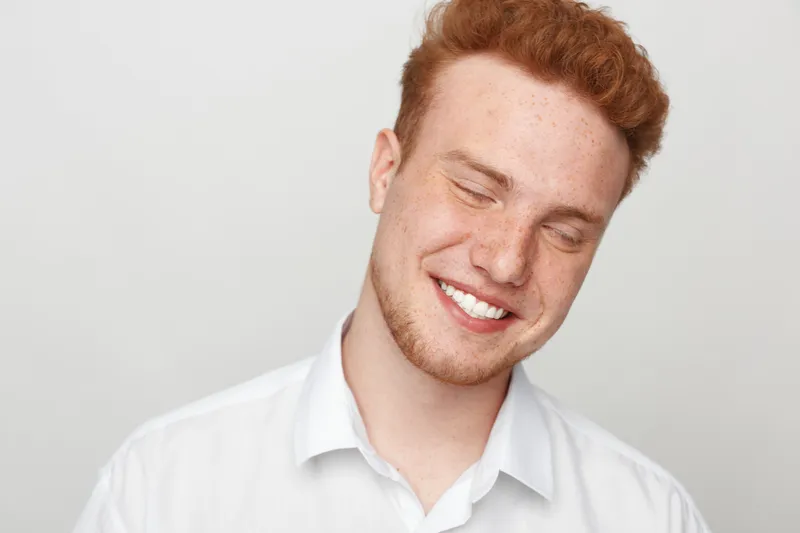 Smiling young man with red hair and freckles wearing white shirt closes eyes and shows genuine happiness. Portrait on light background. Concept of joy, youth, and natural expression.
