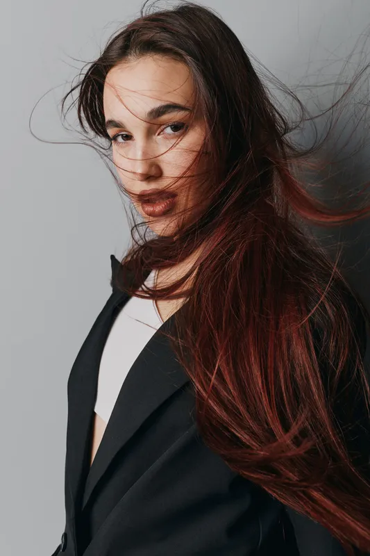 Young woman with long flowing hair wearing black blazer and white top posing against gray background for fashion portrait and beauty concept in studio lighting.