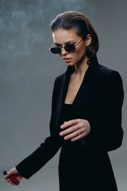 Stylish woman wearing sunglasses in a black blazer with a smoky background and confident pose.