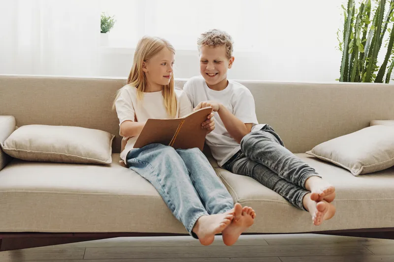 Two happy children sit barefoot on a beige couch, smiling and reading a book together in a bright modern living room. Concept of friendship, learning, and childhood.