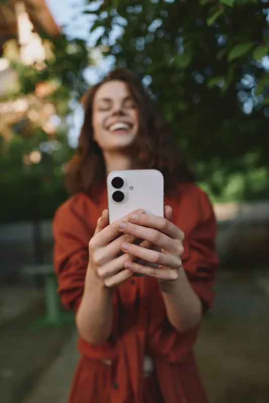 Selfie scene shows a young woman with curly hair smiling as she holds a smartphone in a park, capturing a candid moment outdoors with warm daylight and a relaxed mood