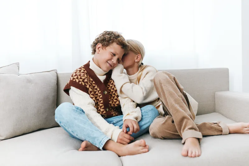 Two young friends relax on a light sofa, sharing a tender moment in cozy sweaters. Soft natural light highlights smiles, warmth, and simple, genuine friendship.