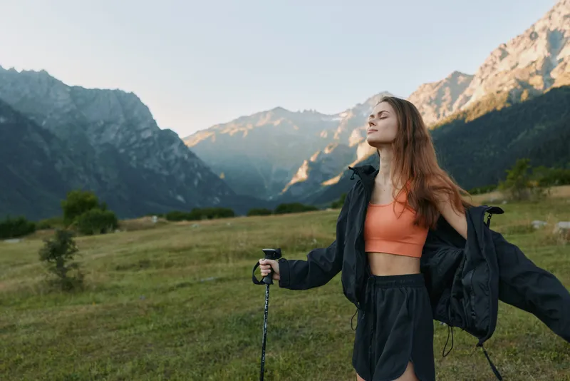 A young woman stands outdoors in a scenic mountain valley, wearing sporty attire and a jacket, looking serene and confident as she poses with hiking poles.