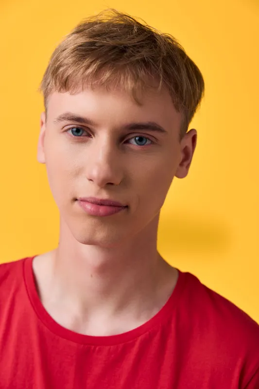 Closeup of a young man with a calm facial expression wearing a red shirt against a bright yellow backdrop, showcasing a clean studio look and modern headshot style.