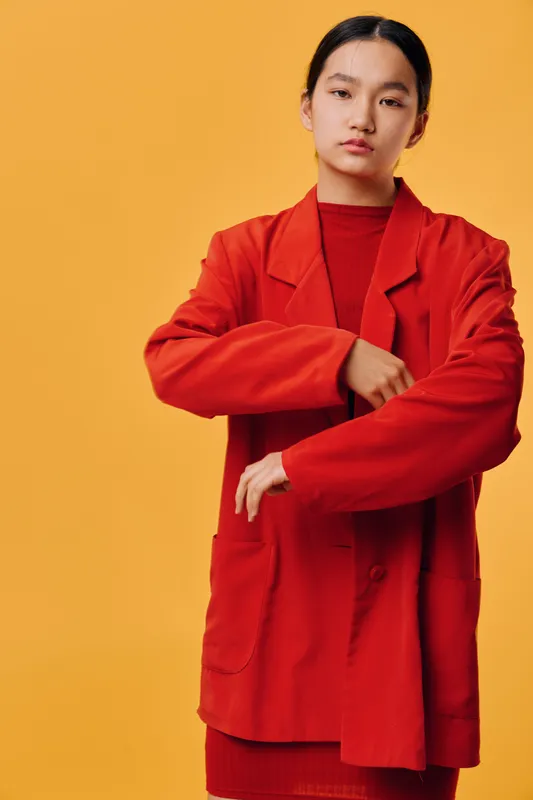 Young woman wearing red jacket and sweater standing in front of orange background, looking confident and adjusting sleeve with serious expression on face.