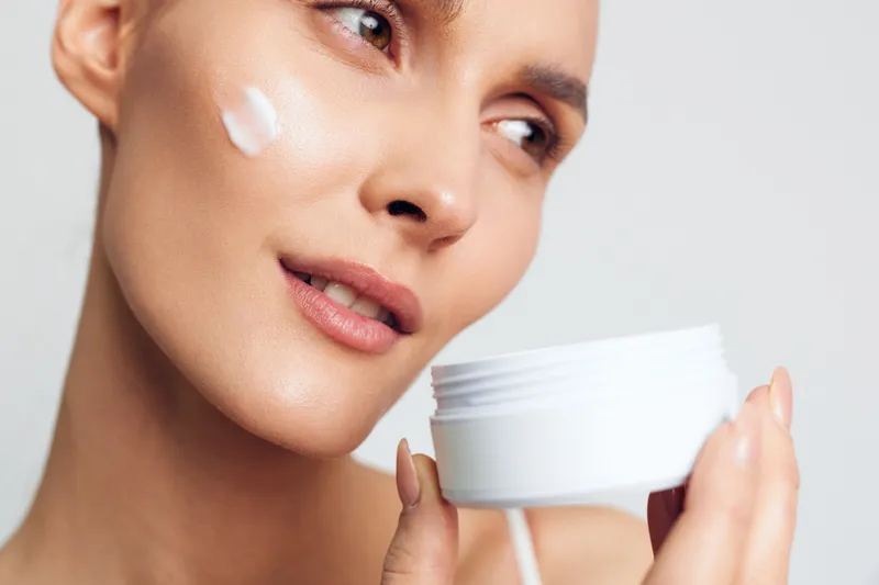 A close-up of a woman with clear skin applying white face cream on her cheek, holding an open jar. The image represents skincare routine and beauty care concept.