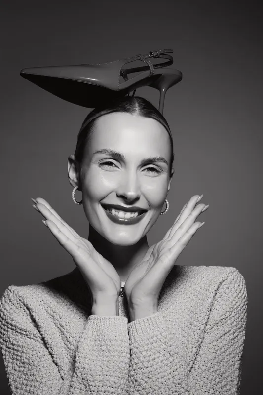 Smiling white woman with fair skin wearing a knitted sweater poses with high heel shoes on her head in a creative studio portrait. Concept for fashion, humor, and modern style.