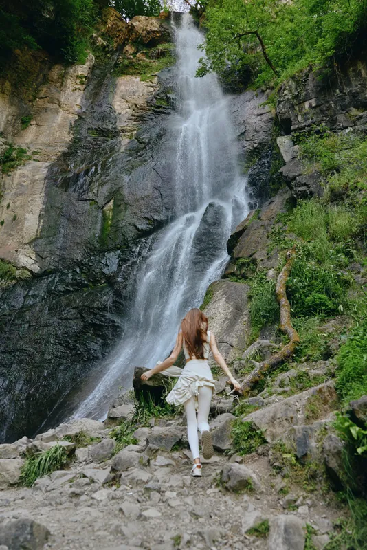 woman hiking near waterfall in lush green forest, wearing white outfit, natural landscape with rocky terrain, adventurous and serene atmosphere
